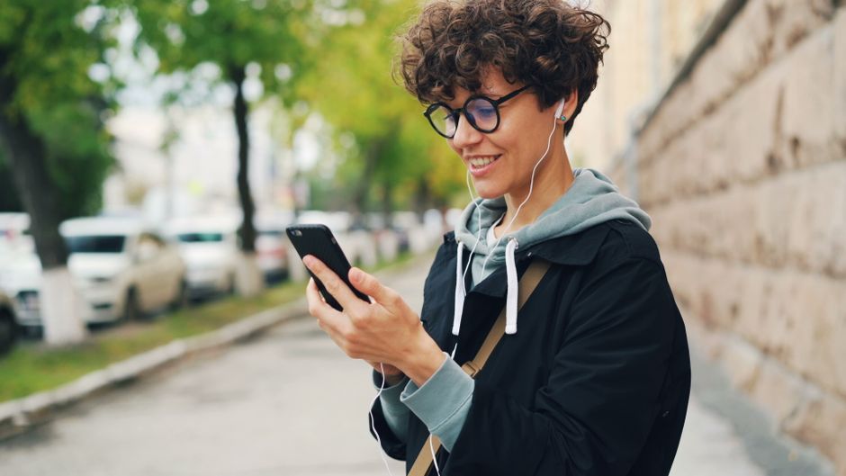 A woman walking down a city street, earphones in, smiling at her phone